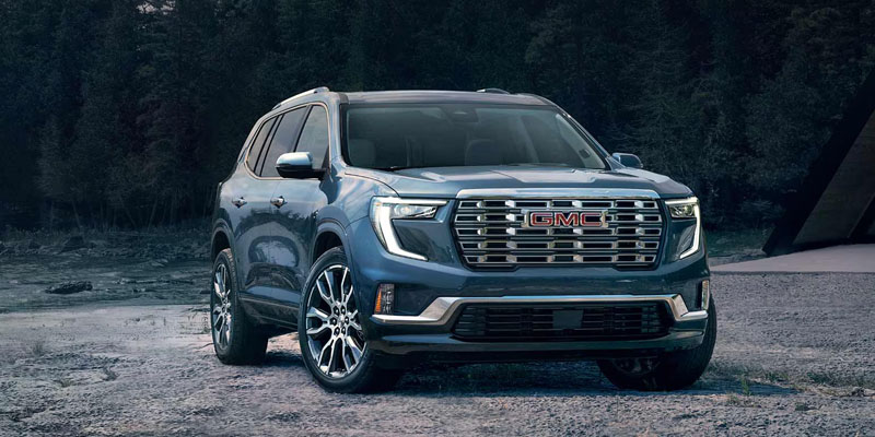 A dusty blue 2026 GMC Acadia SUV is parked on a rough grey road in the mountains.