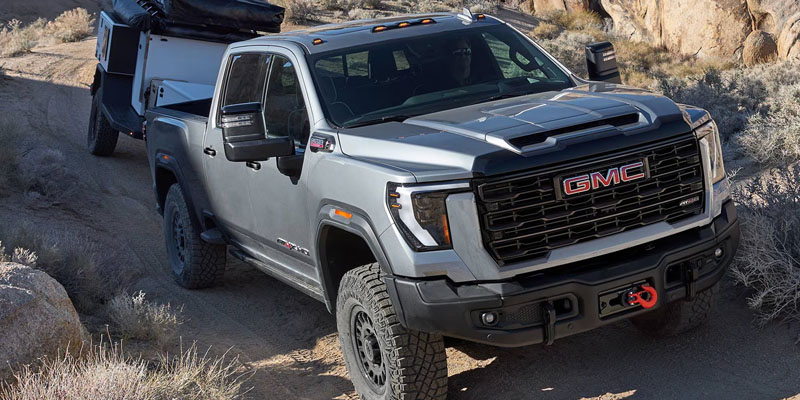 A close-up view of a grey 2026 GMC Sierra 2500 truck is hauling a small 2-wheeled trailer up a very rough dirt road in a dry desert mountain area.