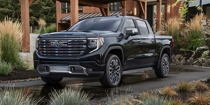 A shining black 2026 GMC Sierra 1500 SUV is parked on a blacktop carport in front of a chic desert home, surrounded with a garden of appropriate plants.