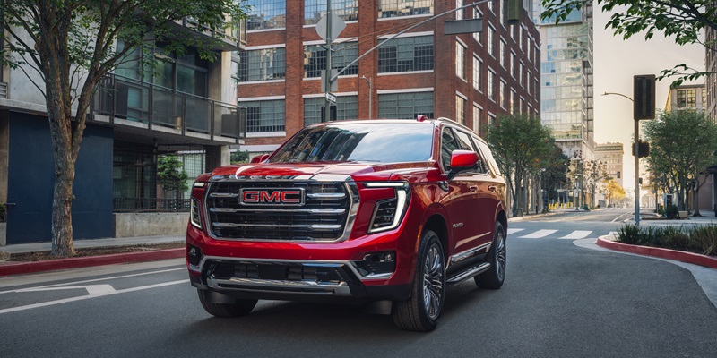 A shining red 2026 GMC Yukon SUV is being driven toward us on a street in the middle of the business district of a small city.