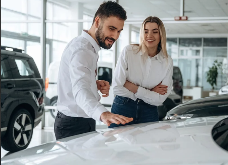 White-shirted male and blonde female are admiring a white GMC SUV with smiles and a hand-pat as they admire the vehicles for sale in the bright white sales room full of vehicles for sale.