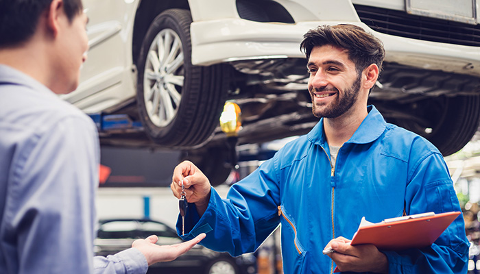 A certified mechanic at a GMC Service Center is handing the keys to the owner of the vehicle lifted above which he has just finished checking out.